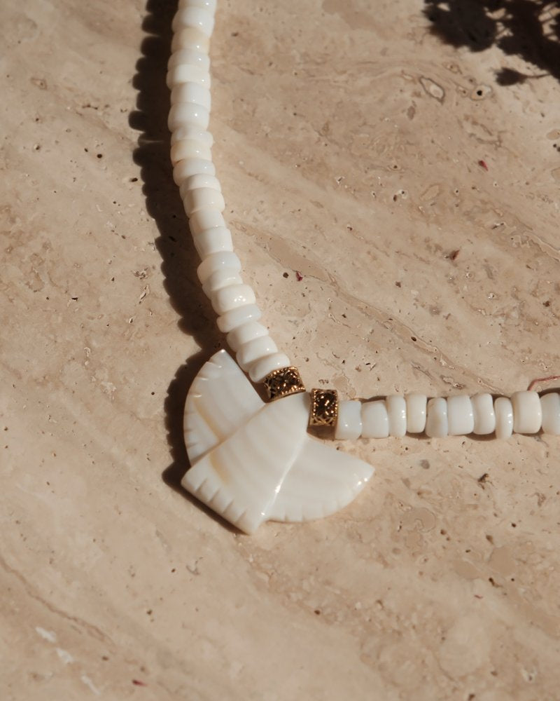 Collier en perles blanches et pendentif en forme d’oiseau, présenté sur une dalle en pierre beige